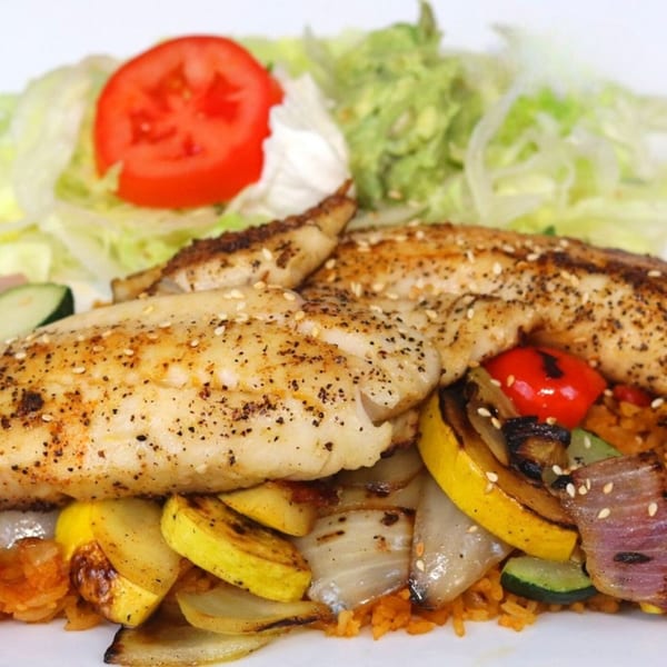 Grilled fish fillets with seasoned vegetables, rice, and a side of lettuce, tomato, and guacamole.